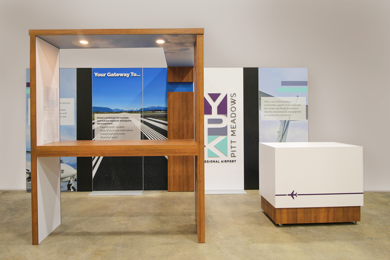 10x20_modular_YPK modern H-line trade show exhibit for an airline. A back wall showcases a geometric logo and airport runway with mountains in the distance. A wooden canopy connects to a counter at the front of the display. The materials are white, and wooden veneer with sky decals on the canopy ceiling. Pot lights are built into the ceiling canopy above the counter area. Another rolling counter is featured with a cutout of an airport runway and plane.