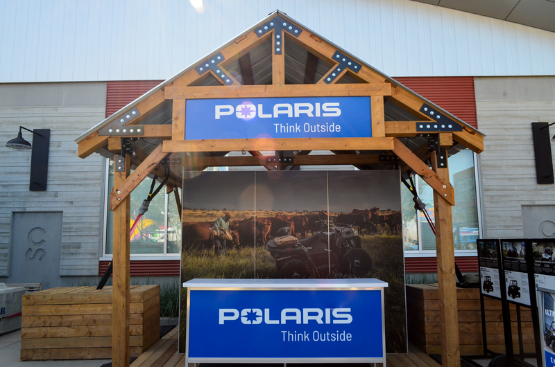 custom fabrication outdoor exhibit. Wooden structure with a tin roof, branded counter, and header sign. the backdrop graphics show a Canadian ranch where cattle are being herded across the prairies