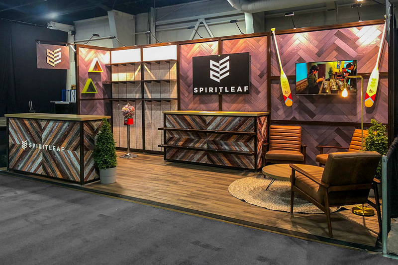 10x20 linear modular trade show booth for a Cannabis retailer. Wooden floors fill the booth space with textured herringbone wooden wall panels. Multi-coloured wooden counters in triangular slat patterns are displayed in the middle and to the left. The back wall includes mountain trainglura decor on the walls, as well as retail shelves. To the right is a monitor and paddle fixed to the wall. In front of it is a cozy seating area with two brown recliners, a circular coffee table, and shag area rug.