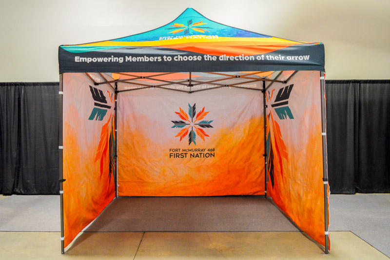 10x10 branded outdoor fabric event tent with double-sided polyester walls and overhead canopy. The tent features a bright orange gradient pattern at the bottom that fades up into solid white near the top of the fabric walls. Multiple arrow tails form a circular logo for Fort McMurray First Nation. The top of canopy near the peak turns into a vibrant turquoise. The fabric walls are secured to each other and the walls with zippers and velcro straps.