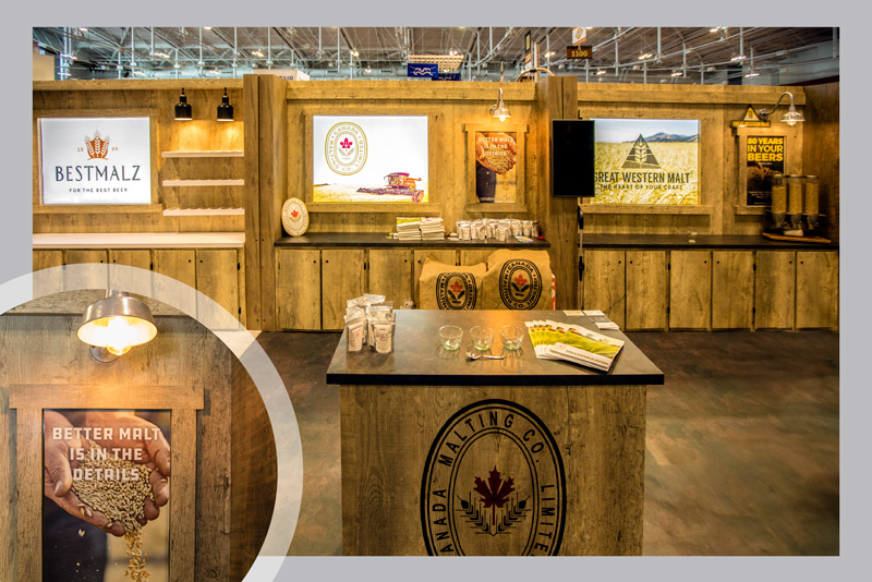 10x30 linear trade show booth for Canada Malting Co at the Brewers Association annual event. The booth features a rustic wooden material with lower cabinets for storage. The walls above feature backlit graphic panels that showcase Canadian fields and barley farming. There is also custom overhead lighting and shelving for displaying products and samples.