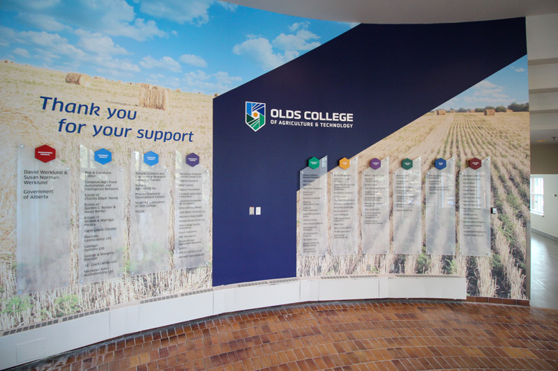 Albertan post-secondary donor recognition wall for the Olds College of Agriculture and technology. A geometric background is shown with blue paint and an Albertan prairie vinyl wall graphic that illustrates a field. The college name and logo is a dimensional acrylic display. Multiple frosted acrylic rectangular panels showcase the names of donors.