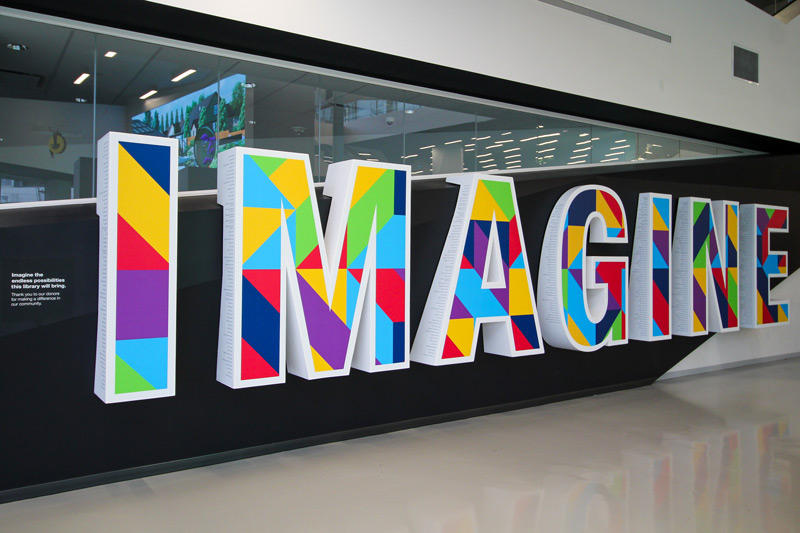 Large and colourful 3D letters built into a library wall that spells "Imagine". The front of the letters have colourful geometric vinyl, and the sides of the letters have the names of donors printed on the sides. This is a unique donor recognition monument for the central Edmonton Public Library in Alberta, Canada.