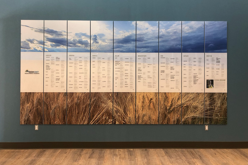 Modular Vulcan County donor recognition wall in Alberta. 9 rectangular panels showcase donor names in front of a prairie scene graphic. Dramatic blue clouds are shown above the names, white a wheat field is shown below as part of one large image.