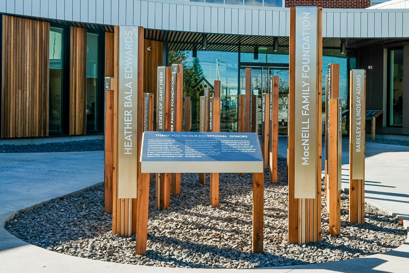 Outdoor donor recognition monument for the Calgary Humane Society in Calgary, Alberta. An angled rectangular panel is at the front of the monument outlining the organization. Within a rock bed, the rest of the monument is made from rectangular cedar posts layered with etched stainless steel panels that recognize donor names.
