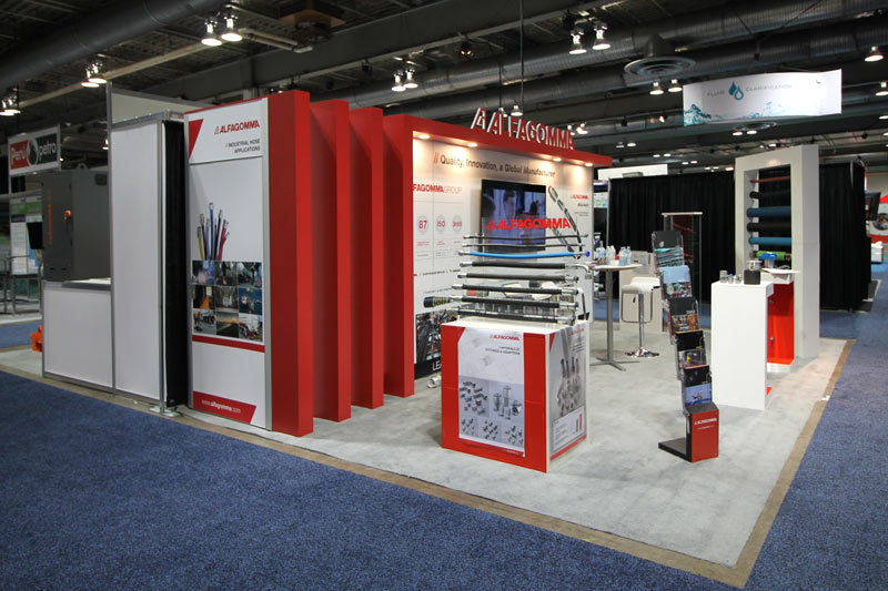 photo on a left angle of the 20x20 trade show display. underneath of the extended red pillars is a branded wall graphic, which serves as a door leading to a storage room. A small box-shaped branded counter showcases products, with a seating area in the middle of the open-concept booth. 