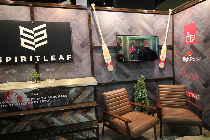 corner seating area for trade show booth clients. Two comfortable brown leather chairs surround a coffee table. Behind them is a wall-mounted monitor between to mounted canoe panels. Graphic decals are mounted to the wall on the right. A neutral wooden herringbone pattern is used for the decorative wall material. 