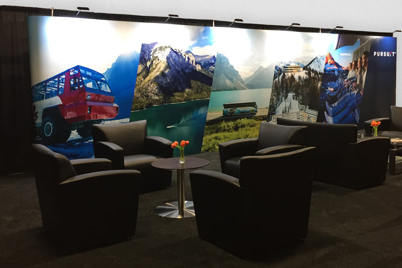 comfortable black armchairs surrounding a coffee table with other couch seating off to the right of the photo. A colourful 30ft portable trade show display is in the background showing vivid imagery of the Canadian Rocky Mountains, illuminated by overhead lights.  