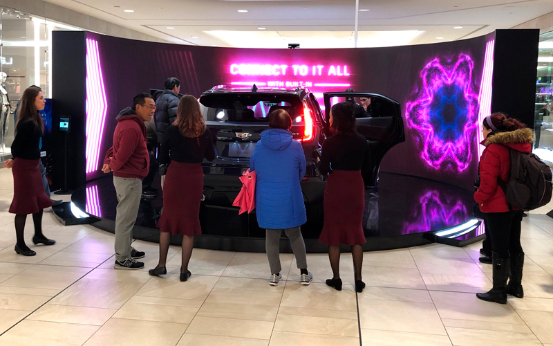 marketing display for Cadillac inside a Montreal shopping mal. The vehicle sits on an elevated black oval platform, facing a curved video wall display with colourful graphics. 