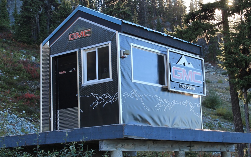 construction of a branded exterior ski race hut on the side of the Whistler mountain. The walled tructure with an A-frame style roof is about 10 feet long and 8 feet wide. A single window and door are on the left side of the display, while the front features a larger window next to a dimensional branded sign. 