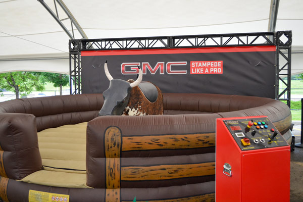 mechanical bull outdoor display during the Calgary Stampede. The bull pen is inflatable with graphics that make it look like an enclosed pen. A branded "GMC" truss sign is behind the setup, with a control station for the bull to the right.