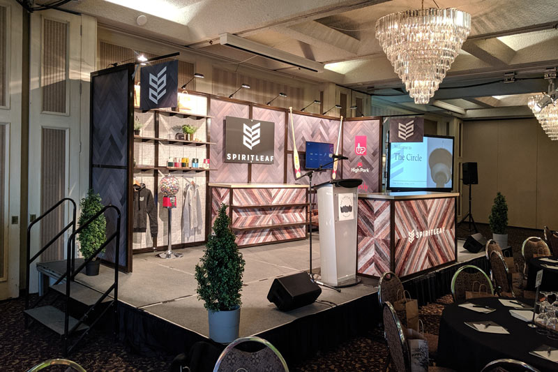 10x20 booth set up on a stage for a custom AGM display. A portable podium has been added to the front, along with live plants and stairs leading up to the stage.
