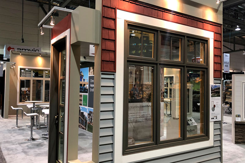 close up of a freestanding wall display on the corner of a trade show booth. A window is showcased with stained brown accents, with red siding panels at the top, and grey siding below. 