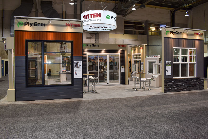 Custom trade show exhibit with freestanding walls showcasing exterior building products such as siding, as well as windows and doors. A cylinder-shaped overhead handing sign is lit up to showcase the Mitten & Plygem logo. The open-concept booth features bar stool seating in the middle, overhead singage and lighting on the freestanding wall displays, and rental carpet. This booth was featured at the WRLA trade show. 
