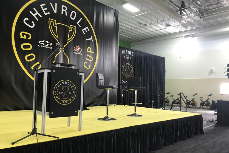 branded event stage for the Chevrolet Good Deeds Cup. A raised stage features a vinyl backdrop, fabric banners, and furniture in black. An Octanorm podium with the logo is also featured to the left of the stage.