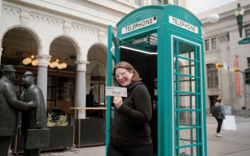 Winner of a free flight voucher to London smiling and holding up their voucher in front of a custom-fabricated branded phone booth in Calgary, Alberta