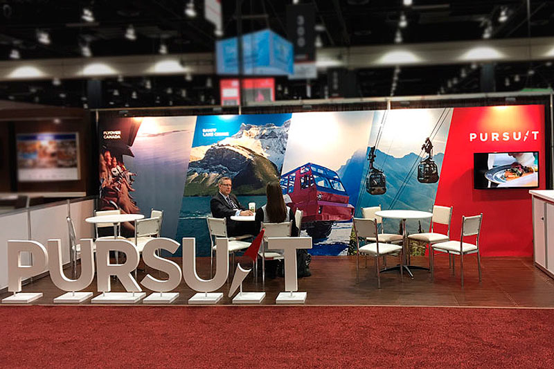 10x30 pop-up display for a Canadian and Worldwide tourism company. The backdrop features scenes throughout the Canadian rockies, with a tv monitored to the backdrop on the right. White rental furniture is setup up in the booth, along with freestanding 3ft-tall letters of the company name set up in front to the left of the booth. 