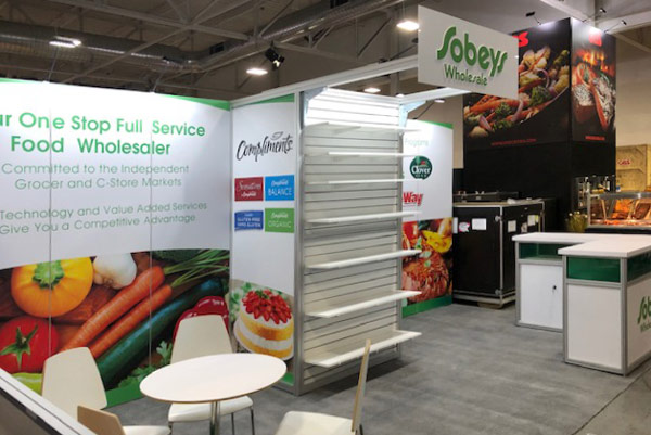 10x20 rental trade show booth for Sobey's groceries. The middle section extends outward with shelving to showcase retail products. At the top of the shelf extending towards the front of the booth is a sign panel with dimensional letters. The right of the booth has an "L" shaped octanorm reception counter, and on the right is rental chairs and table for a meeting area.