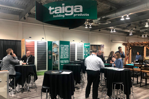 trade show display in calgary with custom building materials showcased on a beMatrix branded wall display. A triangular geometric fabric sign is rigged from the ceiling and showcases the brand's logo. Circular bar chairs and stools are set up in front of the display with black table cloths.