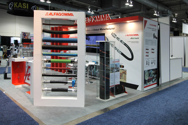 right side of the 10x20 modular booth which features a freestanding rectangular product display. A white thick frame 4ft wide by 8ft tall showcases 10 different hoses help up by horizontal rods running through them. A glass case on the right of the dispay showcases a large black coiled hose about 1ft in width and 5 feet tall. Next to the enclosed glass case is a freestanding graphic panel outlining product specs and details. 