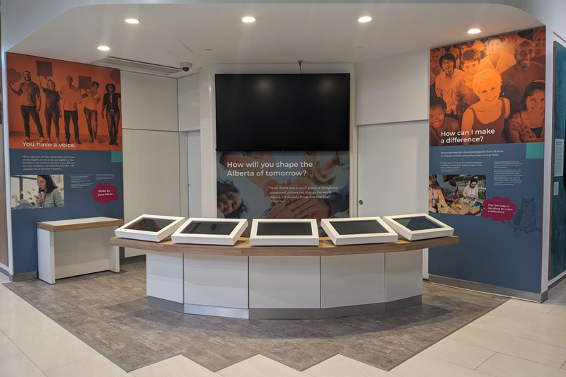interactive digital exhibit display custom built in Edmonton, Alberta. 5 touchscreens are on display at waist height. Graphic wall displays and a wall-mounted tv screen are in the background of this corner exhibit. 