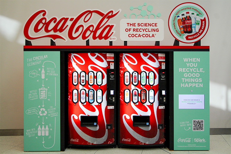 two pop vending machines features in an inclosed display with vinyl graphics and a touch screen. Extended overhead signage is mounted to the top of the display with the Coca Cola Logo and other dimensional signage.