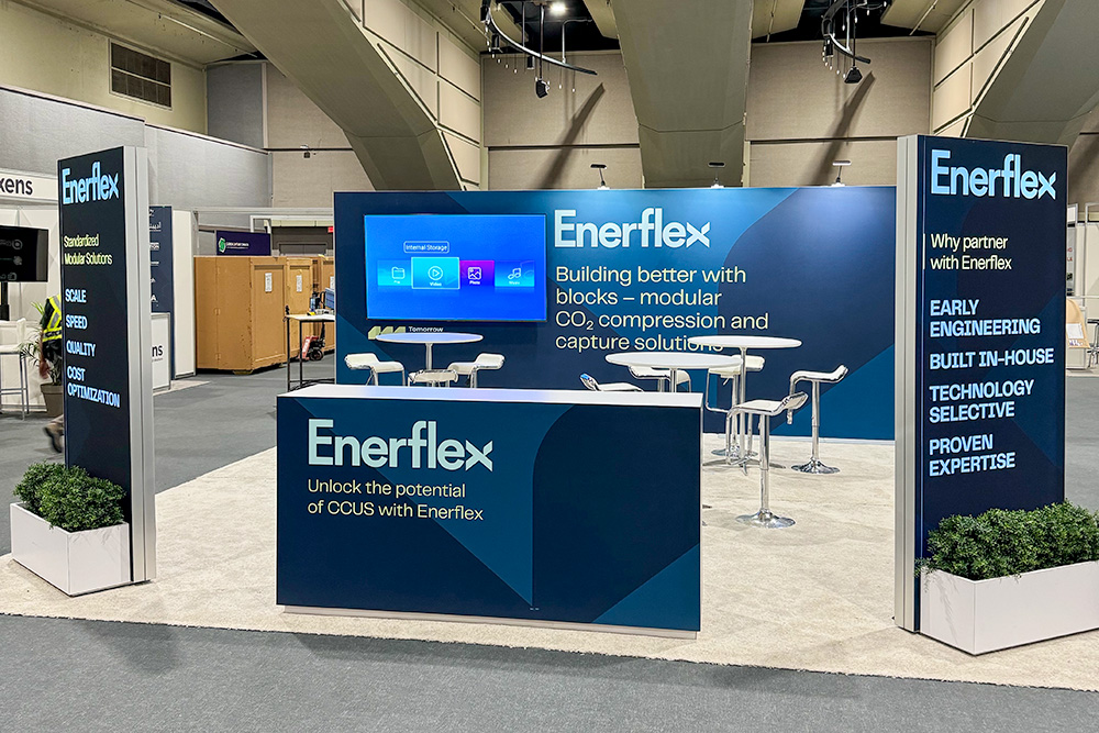 20x20 rental trade show booth in Calgary, Alberta. The back wall features a wall-mounted monitor on the left. The middle has rental furniture for seating. The front of the exhibit features a branded counter and two freestanding wall towers with planter boxes placed in front.