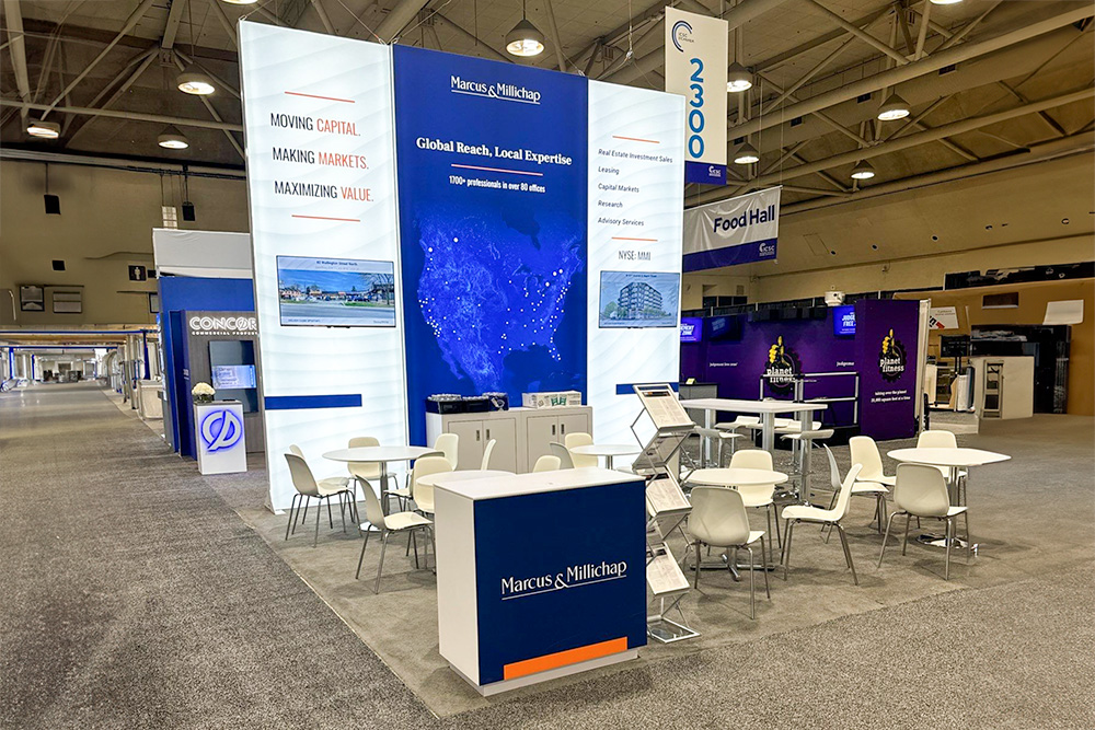 20x20 island trade show booth rental for a trade show event at the calgary BMO centre in Alberta. White rental chairs and tables and a branded counter are placed in front of a backlit fabric display. 