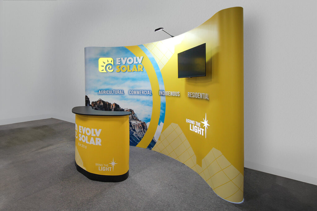 backlit pop-up display for a solar company in Alberta. Imagery of the Rocky Mountains is shown, with a monitor mounted to the display on the right. In front is a boomerang-shaped portable podium with a branded panel. 