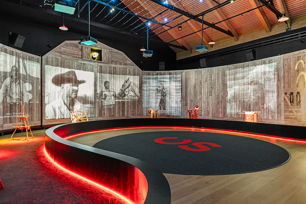 horseshoe-shaped multimedia presentation area in Calgary. The backwalls are made from boards designed to give the shape of a rustic barn for the backdrop Multiple images are screened onto the backdrop showing historic rodeo and ranching footage in alberta. The horseshoe permiter features red LED lights at the flor, which illuminate ground designed to look like dirt on the rodeo track. A variety of historic objects are featured in this area, highlighted with spotlights. 