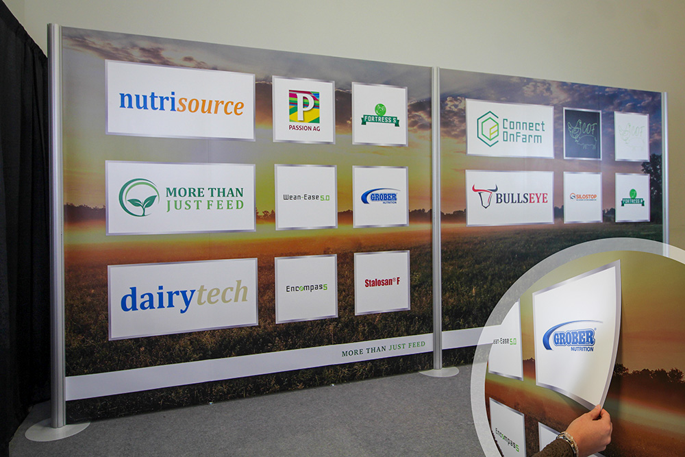 10x20 modular trade show display for an Albertan agriculture business showing a field in the background. square and rectangular magnetic decals are placed on the panels, showing a variety of brands that they work with. The inset image on the right shows a close up of the magnetic decal being peeled off. 