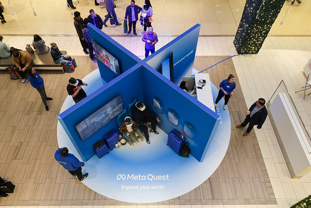 birds-eye view of a 10x10 "x"-shaped free standing wall with 4 sections. This sits atop of a blue circular floor decal with Meta branding. Each section has users wearing a VR headset and setting out different environments related to travel, working out, and gaming.