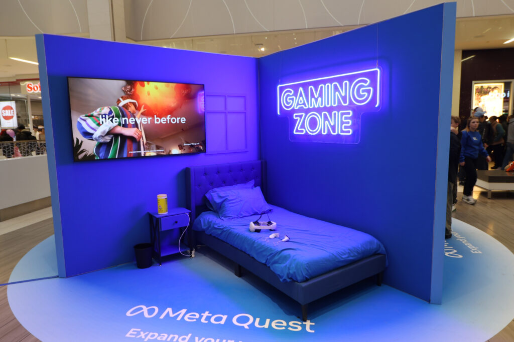 corner section of an open free-standing display at West Edmonton Mall. A blue room has a single bed in the corner, with a custom "gaming zone" LED sign on the right wall. On the left is a tv monitor advertising the Meta Quest VR headset, which sits on the bed waiting for users to test it out. 